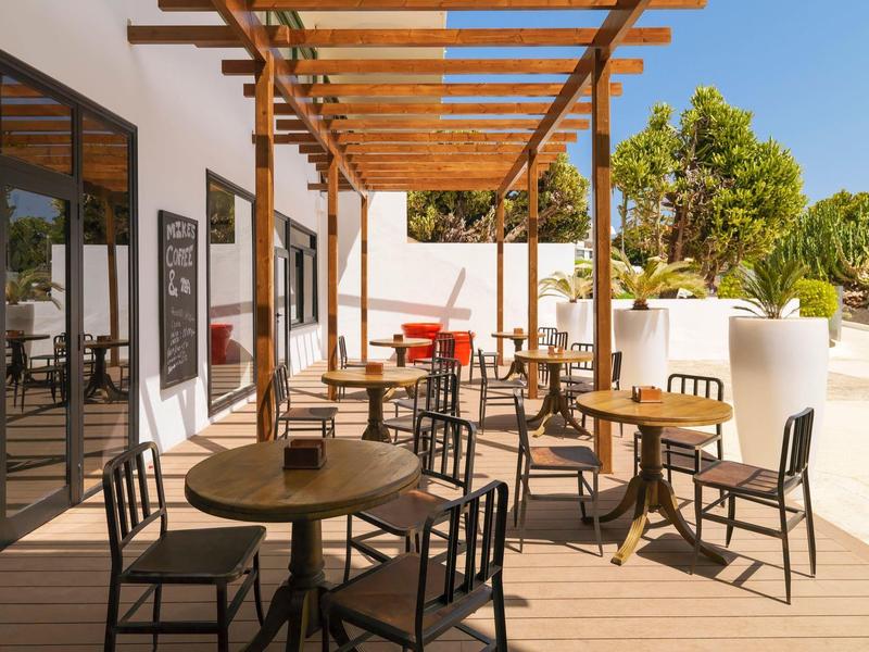 Außenbereich mit Holzterrasse, Tischen, Stühlen und Holzpergola neben Glasfenstern und grünen Pflanzen.