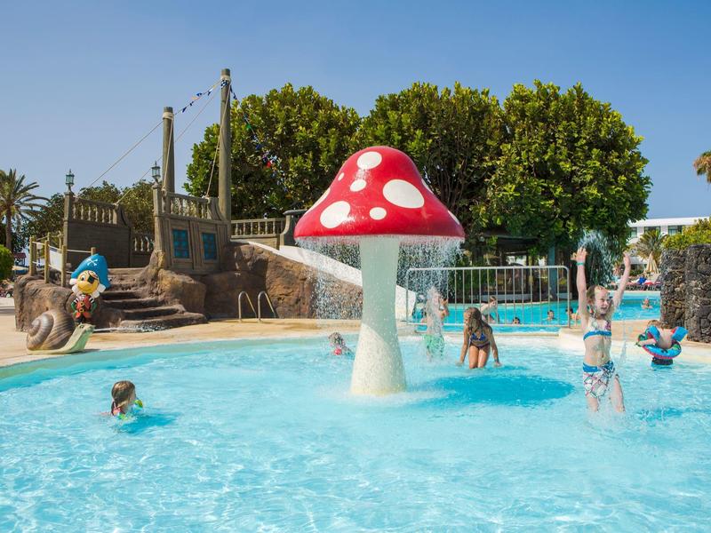 Kinder plantschen im Pool mit großem roten Pilzbrunnen und blauem Himmel im Hintergrund.