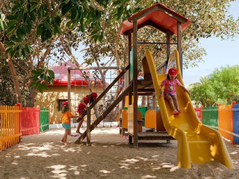 Kind spielt auf gelber Rutsche in buntem Spielplatz mit Baum und Sand unter dem Spielgerät.