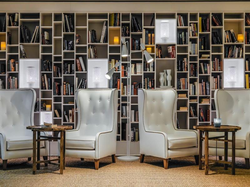 Vier helle, bequeme Sessel mit kleinen Holztischen vor einer großen Bücherwand mit vielen Büchern.