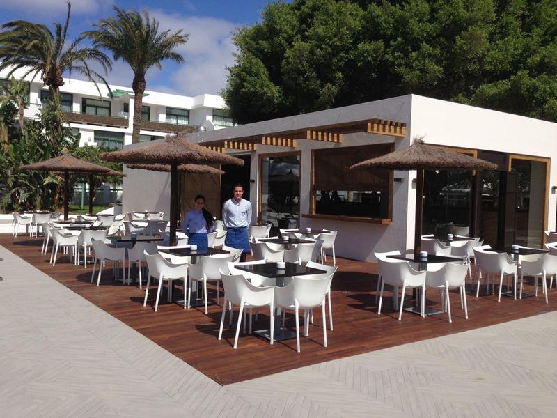 Außenbereich eines modernen Cafés mit weißen Stühlen und Tischen auf Holzterrasse unter Palmen.