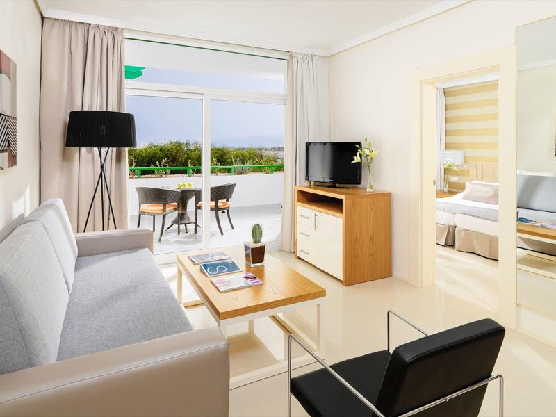 Helles Wohnzimmer mit hellgrauem Sofa, schwarzem Sessel, TV, Tisch und Blick auf Balkon mit See.