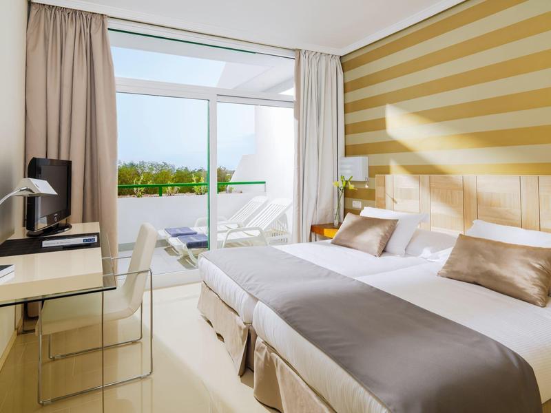 Hotelzimmer mit zwei Einzelbetten, Schreibtisch, Stuhl, Balkon mit Glasgeländer und gelb gestreifter Wand.