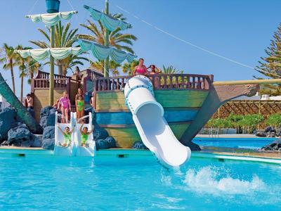 Scivoli d'acqua in piscina di un resort soleggiato con palme e persone che si divertono.