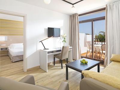 Bright hotel room with workspace, sofa, and balcony overlooking the sea.