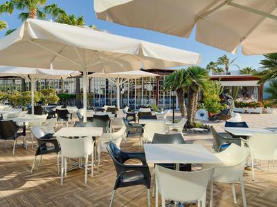 Freiluftrestaurant mit weißen und schwarzen Stühlen, Tischen und großen Sonnenschirmen bei Sonnenschein.