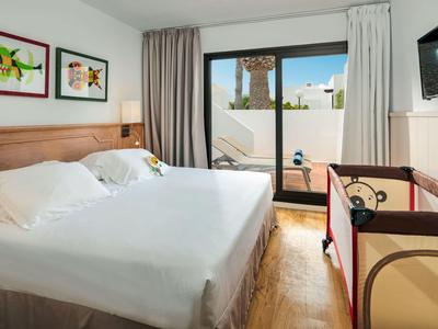 Bright hotel room with double bed, crib, and terrace access overlooking a pool.