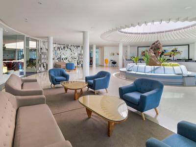 Luminosa hall dell'hotel con poltrone blu, divani beige e vaso centrale sotto lucernario.