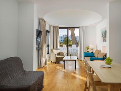 Bright hotel room with wooden floor, sofa, dining table, and balcony overlooking buildings.