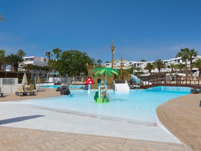 Leisure pool with water features and sun loungers at a hotel resort under clear sky.