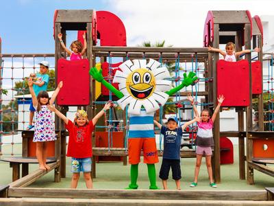 Bambini che giocano su un parco giochi colorato con una mascotte del sole sorridente al centro.