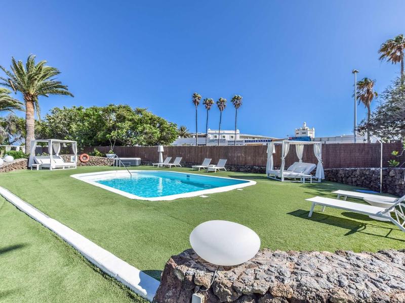Schwimmbad mit blauem Wasser, grüner Rasenfläche, Liegestühlen und Palmen bei klarem Himmel.