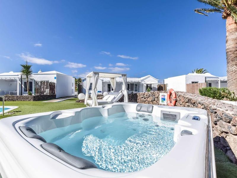 Moderner Whirlpool im Außenbereich mit Pool, Liegen und weißen Bungalows unter blauem Himmel.