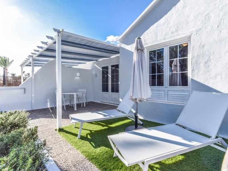 Haus mit weißer Fassade, Terrasse mit Pergola, Tisch mit Stühlen und zwei Sonnenliegen mit Sonnenschirm.
