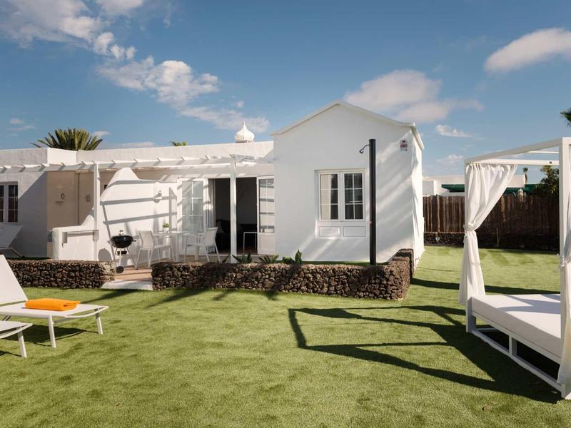 Weißes Ferienhaus mit grüner Wiese, Sonnenliegen und Pavillon unter blauem Himmel.