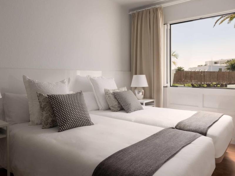 Zwei Einzelbetten mit weißen und grauen Bettwäsche vor einem Fenster mit Vorhängen in einem hellen Zimmer.