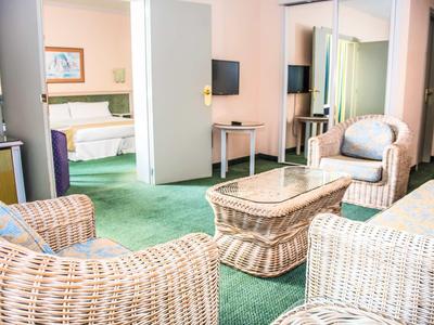 Helles Wohnzimmer mit grünem Teppich, Korbmöbeln und Blick ins Schlafzimmer mit Doppelbett.
