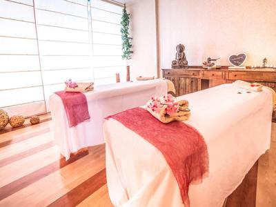 Helles Massagestudio mit zwei Liegen, weißen Tüchern, roten Decken und ruhiger Dekoration.