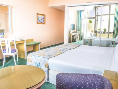 Helles Hotelzimmer mit Doppelbett, Schreibtisch, Stuhl, Sofa, Tisch und Balkon mit Gartenblick.