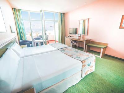 Helles Hotelzimmer mit großem Bett, grünem Teppich, Schreibtisch und Balkon mit Sitzbereich.