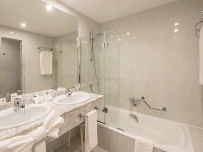 Helles Badezimmer mit Doppelwaschbecken, großer Spiegel und Badewanne mit Glasduschabtrennung.