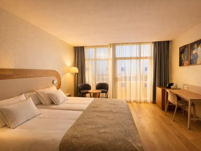 Helles Hotelzimmer mit zwei großen Betten, Holzboden und Sitzecke vor einem Fenster mit Vorhängen.