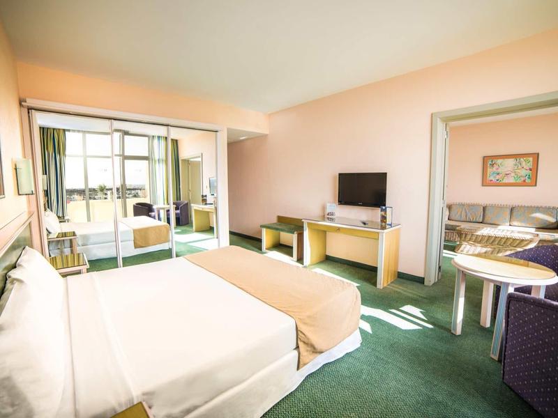 Habitación de hotel luminosa con cama doble, escritorio, televisor y área de sala contigua con sofá.