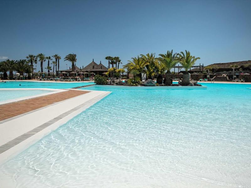 Piscina al aire libre grande con agua cristalina, escaleras blancas y palmeras al fondo bajo cielo azul.