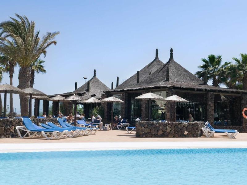 Área de piscina con tumbonas azules, sombrillas y vegetación tropical bajo cielo despejado.