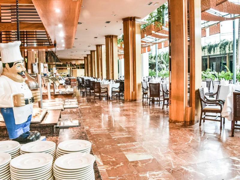 Amplia zona de restaurante con muebles de madera, estatuas y grandes ventanales que proporcionan luz natural.