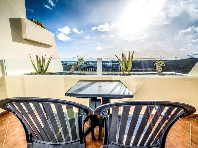 Balcón con dos sillas negras, mesa, plantas y vista a la montaña bajo cielo soleado.