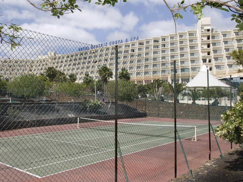 Pista de tenis al aire libre vacía frente a un gran edificio de hotel blanco bajo un cielo nublado.