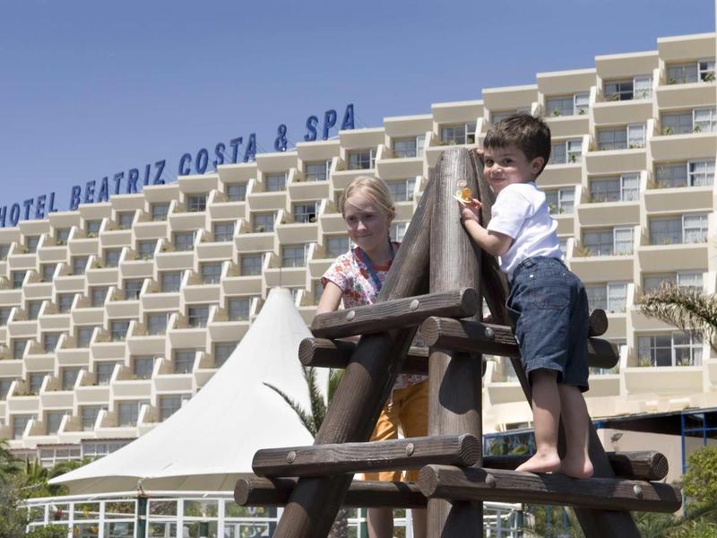 Niños jugando en estructura de madera frente al hotel Beatriz Costa & Spa en un día soleado.