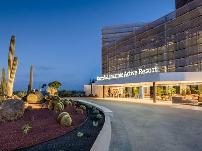 Entrada moderna de hotel con fachada de vidrio y cactus en primer plano al anochecer.