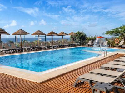 Buitenzwembad met ligstoelen en rieten parasols bij blauwe lucht en zeezicht.