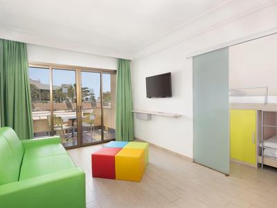 Lichte hotelkamer met groene bank, kleurrijke poef en uitzicht op de stad door het raam.