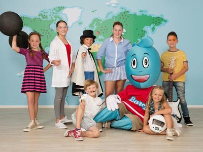Kinderen en volwassenen met een blauwe mascotte voor een wereldkaart binnen.