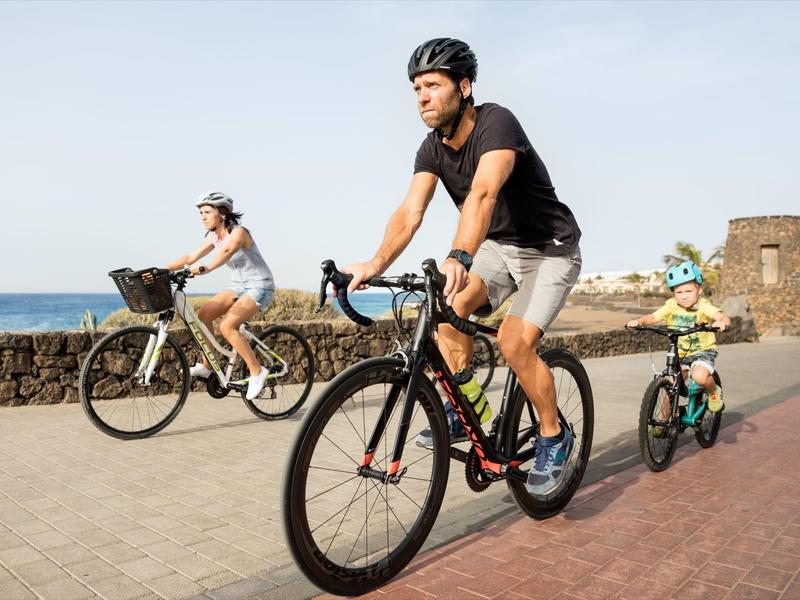 Gezin fietst samen met fietsen en een kinderaanhanger langs een kustpad.
