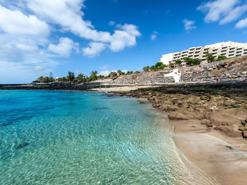 Helder turquoise water bij rotsachtig strand met hotelgebouw en blauwe lucht.