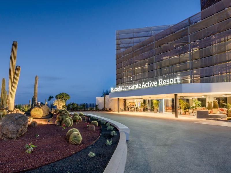 Ingang van een modern resort met glazen gevel en cactussen in de tuin bij schemering.