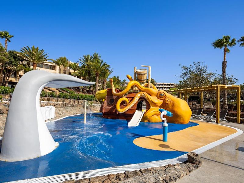 Kleurrijke waterspeeltuin met glijbanen en fonteinen onder een blauwe lucht.