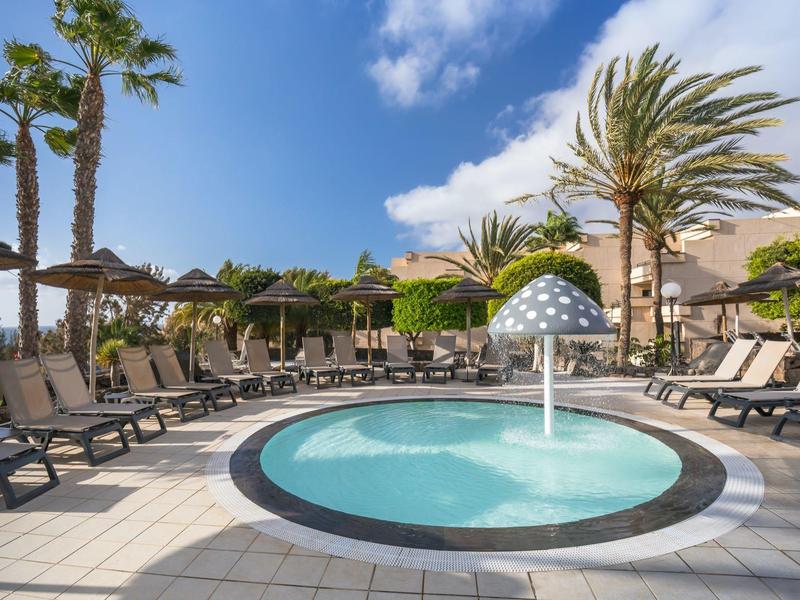 Rond zwembad met paddestoel-waterfunctie en ligstoelen onder palmbomen op zonnige dag.