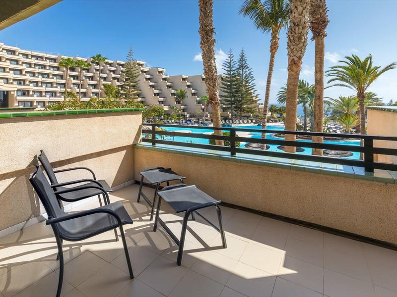 Balkon met drie zwarte stoelen met uitzicht op een zwembad en palmbomen bij zonnig weer.