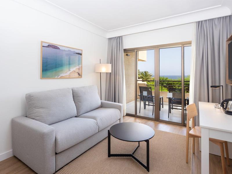 Lichte woonkamer met grijze bank, kleine tafel en balkon met zeezicht.