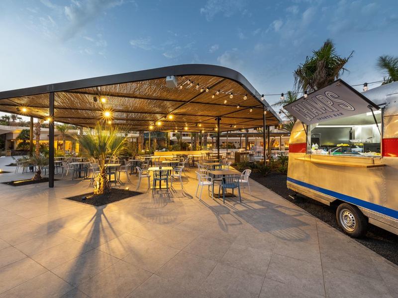 Verlichte openluchtbar met tafels en stoelen naast een foodtruck bij zonsondergang.