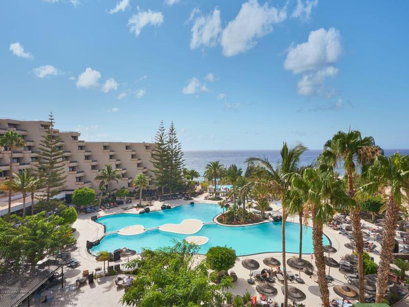 Hotel met groot zwembad, palmbomen en ligstoelen met uitzicht op zee onder een blauwe lucht.