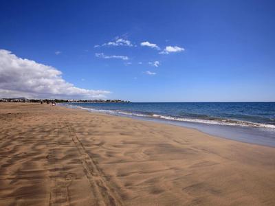 Weite Sandstrand mit Wellen und blauem Himmel, links Wolken am Horizont.