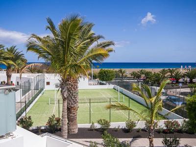 Moderner Tennisplatz mit Palmen, weißem Zaun und Meer im Hintergrund unter blauem Himmel.