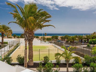 Blick auf einen Tennisplatz mit Palmen und Meer im Hintergrund bei blauem Himmel und Wolken.
