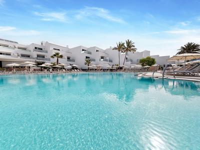 Grande piscina dell'hotel con acqua blu, circondata da lettini e edifici bianchi sotto un cielo azzurro.